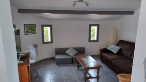 Una sala de estar con un sofá de cuero marrón, una mesa de centro de madera, un sofá gris y una mesita con un ventilador. Hay dos ventanas y un ventilador de techo.