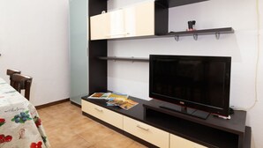 Um quarto com televisão em um suporte, uma mesa com toalha e uma cadeira.