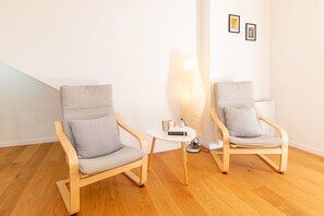 Deux chaises en bois avec des coussins gris, une petite table blanche sur laquelle se trouvent un livre et un vase, et un grand lampadaire.
