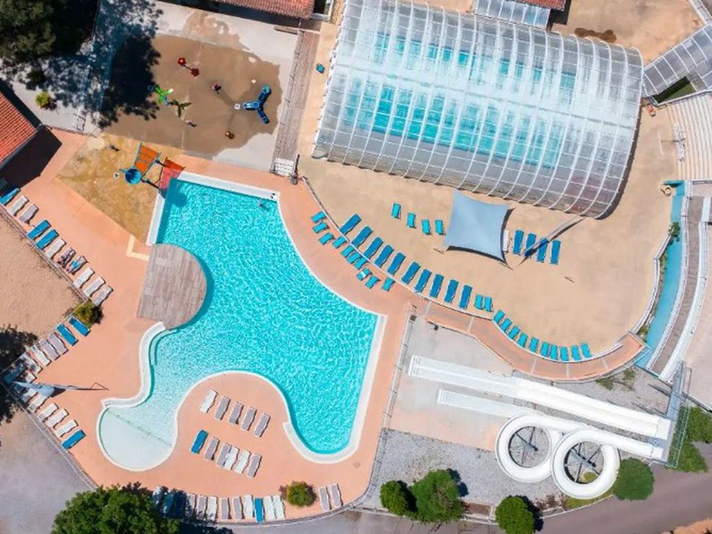 An aerial view of a resort with a large swimming pool, sun loungers, and a greenhouse structure.