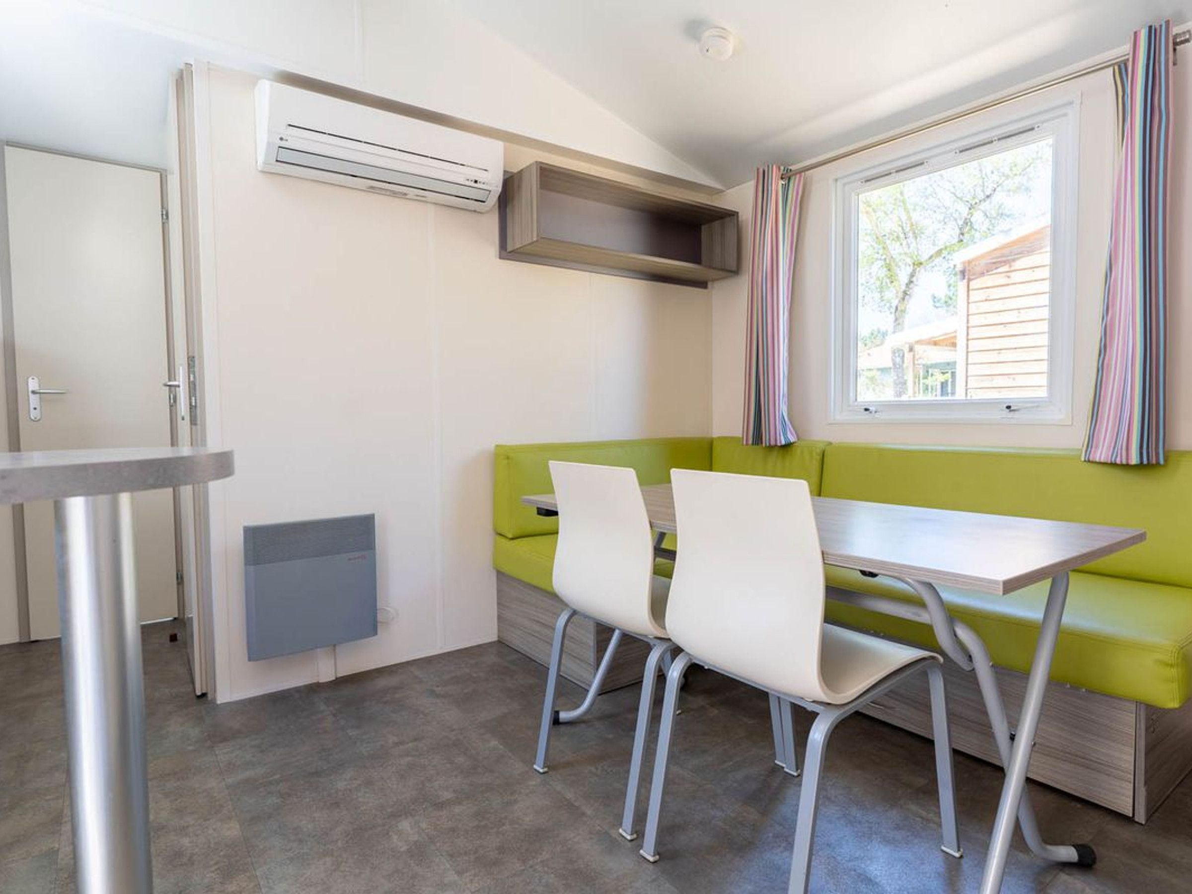 A compact kitchenette with a built-in stove, a window with striped curtains, and a green bench with two chairs.