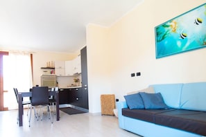 Una sala moderna con un sofá azul, una zona de comedor con sillas, una cocina con armarios blancos y un cuadro en la pared.