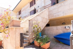 Una escalera de piedra que conduce a la entrada de un edificio, rodeada de plantas en macetas y un objeto azul en el suelo.