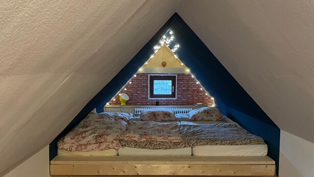 A triangular attic bedroom with a bed, a window, and a brick wall. - Vacation apartment with family bed - ideal for families, groups and fitters (Groß Niendorf)