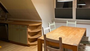 A dining area with a wooden table and chairs, a window with blinds, and a kitchen with a dishwasher. - Vacation apartment with family bed - ideal for families, groups and fitters (Groß Niendorf)