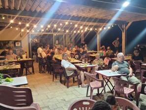 An outdoor dining area with wooden tables and chairs, string lights overhead, and a thatched roof structure. - Mobile Home with Terrace - 6 pers - Comfort in Grand-Serre (Le Grand-Serre)