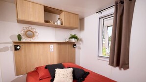 Un dormitorio con colcha roja, cabecera de madera, ventana con cortinas y estante con objetos.