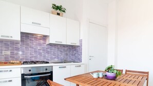 A modern kitchen with white cabinets, a purple-tiled backsplash, a wooden table, and chairs. - Casa Tatti- Calm&Natural (San Giovanni di Sinis)