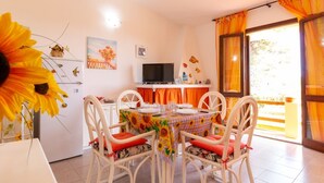 Un comedor con una mesa dispuesta para cuatro, sillas blancas con cojines rojos, un televisor sobre un mueble bajo y un ventanal con cortinas.
