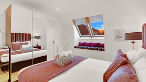A modern bedroom with a large bed, a mirror, a window with a view of the beach, and a wall-mounted shelf with towels. - Old Farm- North East Escapes (Bamburgh)