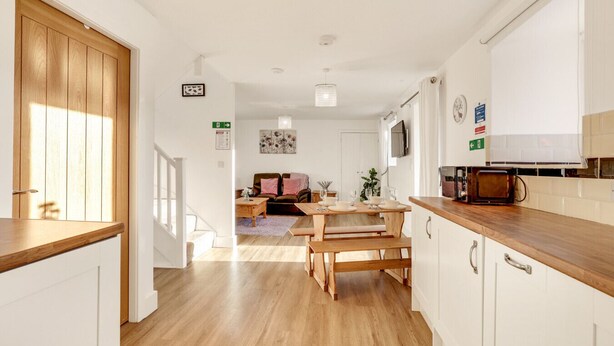 Una cocina moderna con encimeras y armarios de madera, un comedor con mesa y bancos, y una sala de estar con sofá y un cuadro en la pared.