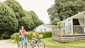 Deux personnes à vélo sur un chemin de gravier, avec une caravane en arrière-plan.