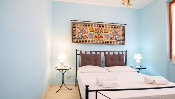 A bedroom with a bed, two bedside tables with lamps, a decorative wall hanging, and a chandelier. - Appartamento Azzurro-Lovely&Quiet (Oristano)