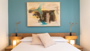A neatly made bed with white linens, a blue wall, and a large abstract painting above it.
