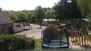 A playground with a trampoline and climbing structures, surrounded by a white picket fence, with houses and greenery in the background.