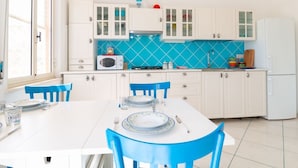 Een keuken met witte kasten, een blauwe betegelde spatwand, een witte tafel met blauwe stoelen en een raam.