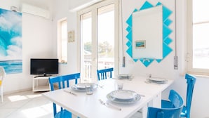 Een lichte kamer met een witte eettafel, blauwe stoelen, een televisie en een groot raam.