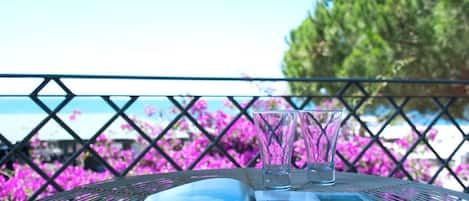 A table with a menu, two glasses, and a view of the sea and pink flowers.