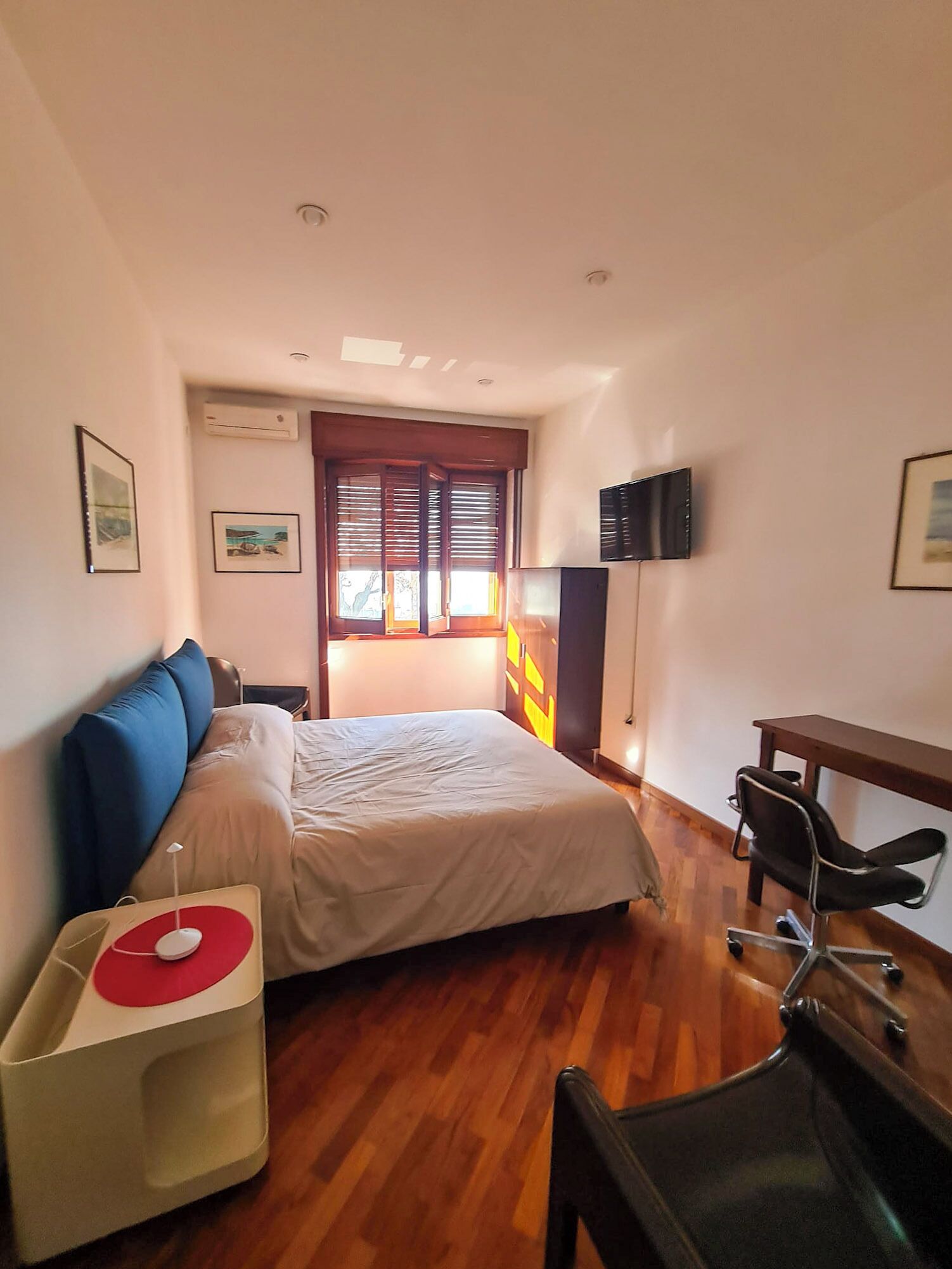 A bedroom with a bed, a desk, a chair, a TV, and a window with blinds.
