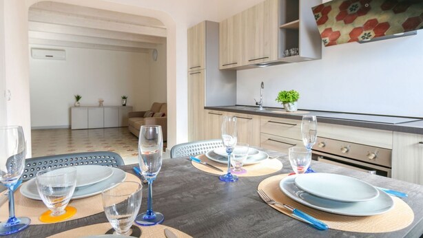 Zona pranzo con tavolo apparecchiato per quattro, cucina con pensili in legno chiaro e un soggiorno sullo sfondo.