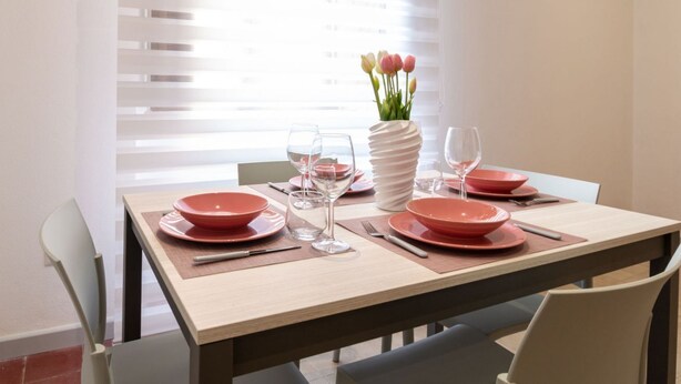 Mesa de jantar posta com pratos vermelhos, copos transparentes e um vaso com flores.