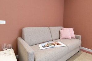 Un sofá beige con un cojín rosa, una revista y dos copas de vino vacías sobre una mesa blanca frente a una pared color melocotón.