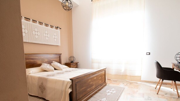 A bedroom with a wooden bed, a bedside table, a chair, a patterned rug, and a large window with curtains. - Cocco e Dessì Home- Angelica (ORISTANO)