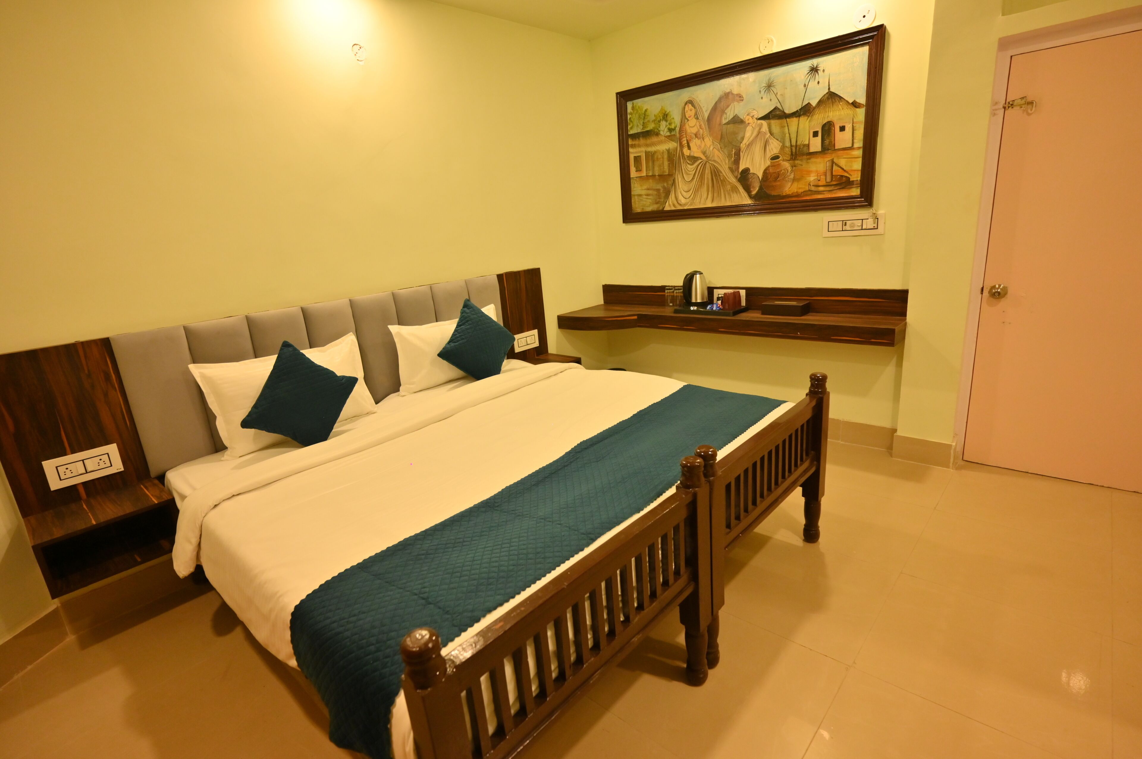 A neatly made bed with a wooden headboard, a teal blanket, and two pillows. There is a painting on the wall above the bed and a shelf with a kettle and a mug below it.