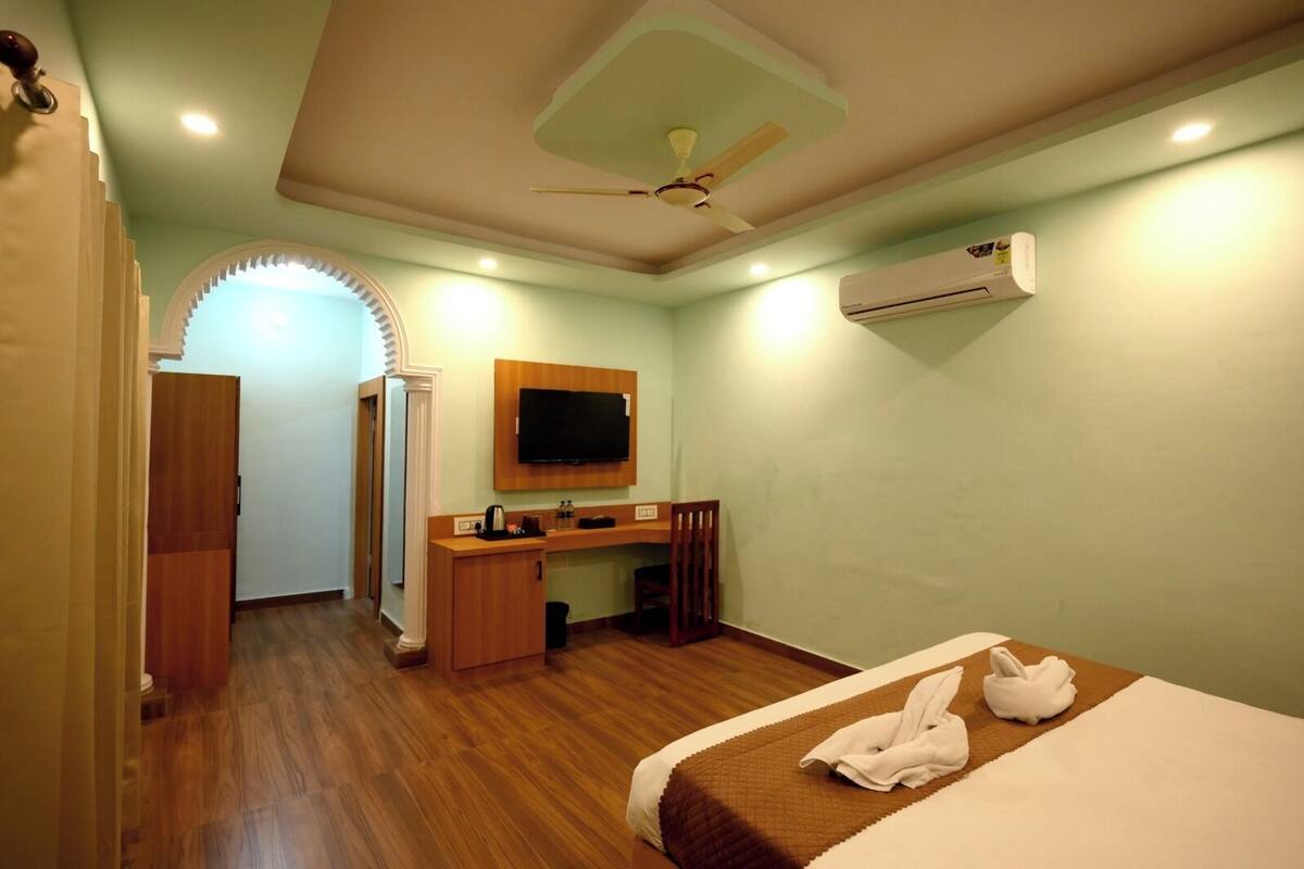 A hotel room with a wooden floor, a bed with a brown bedspread, a desk with a TV, and an air conditioning unit on the wall.