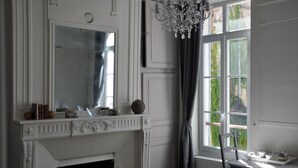 Een kamer met een witte open haard, een grote spiegel, een kroonluchter en een raam met gordijnen.