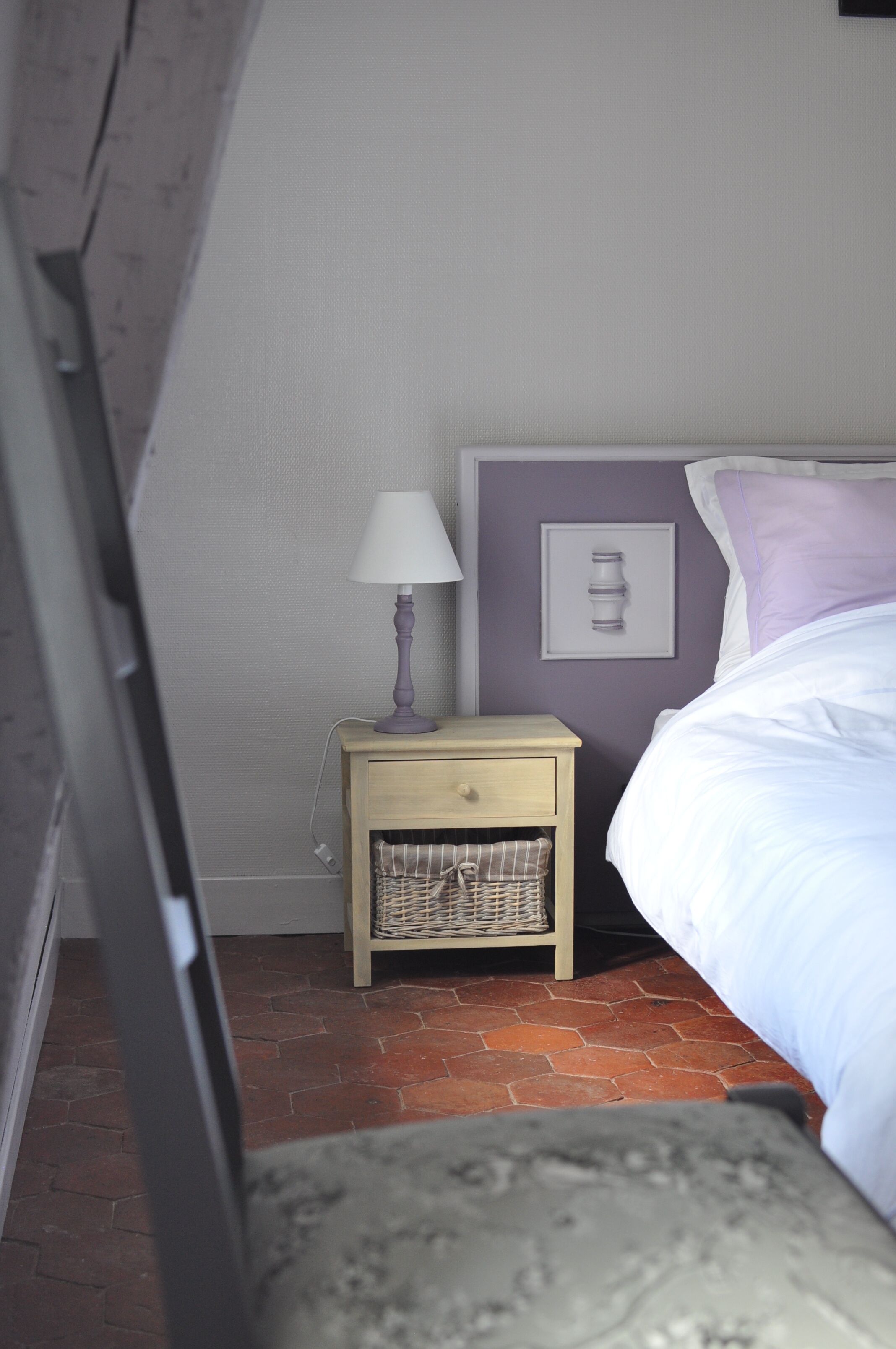 Un dormitorio con cama, mesita de noche con lámpara, interruptor de luz en la pared y una cesta de mimbre debajo de la mesita.