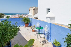 A rooftop terrace with a view of the sea, a castle in the background, and a blue and white building. - Room 'Hostal Santa Catalina 3' with Shared Terrace, Wi-Fi, and Air Conditioning (Conil de la Frontera)