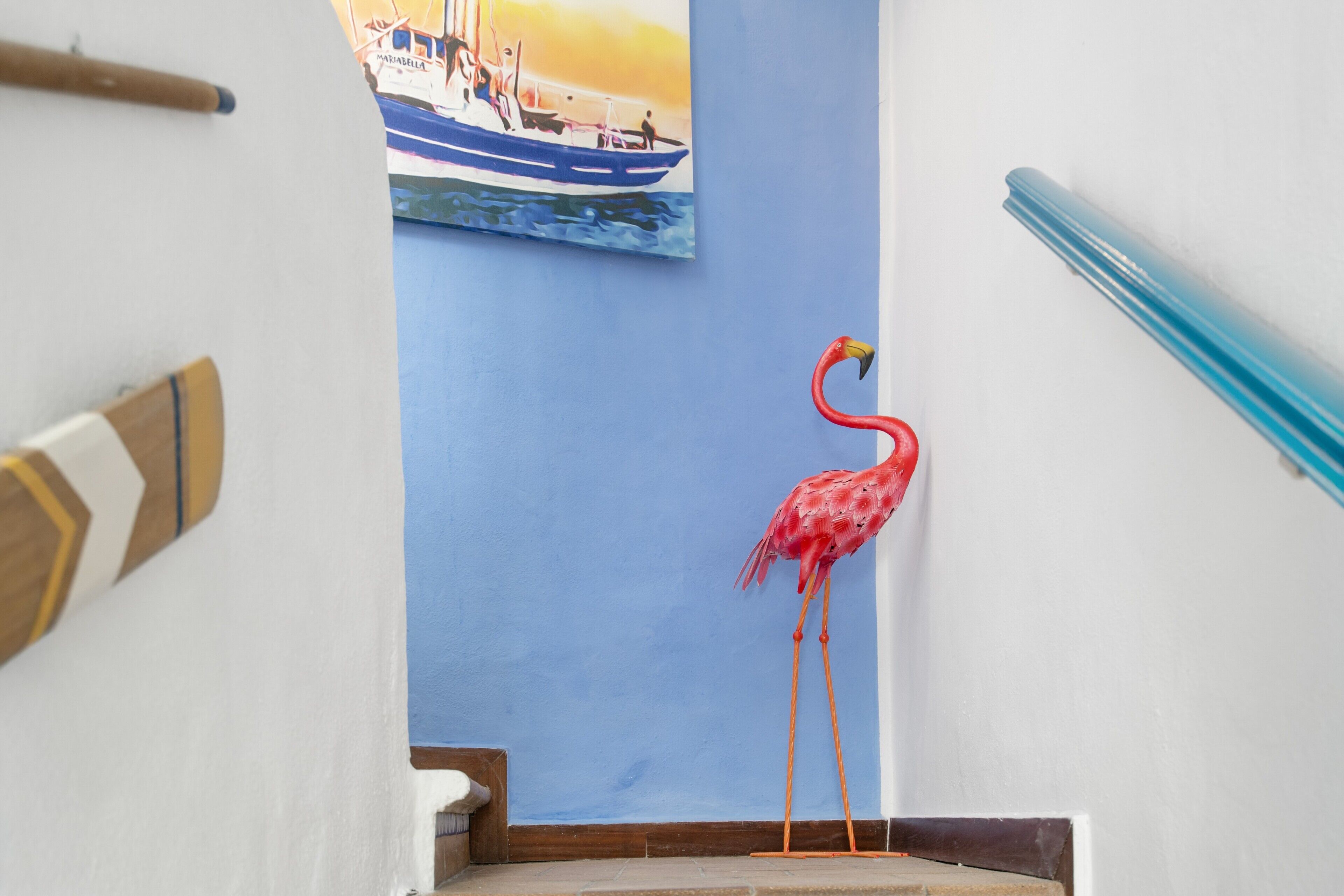 A staircase with a red flamingo statue, a painting of a boat on the wall, and a wooden handrail.