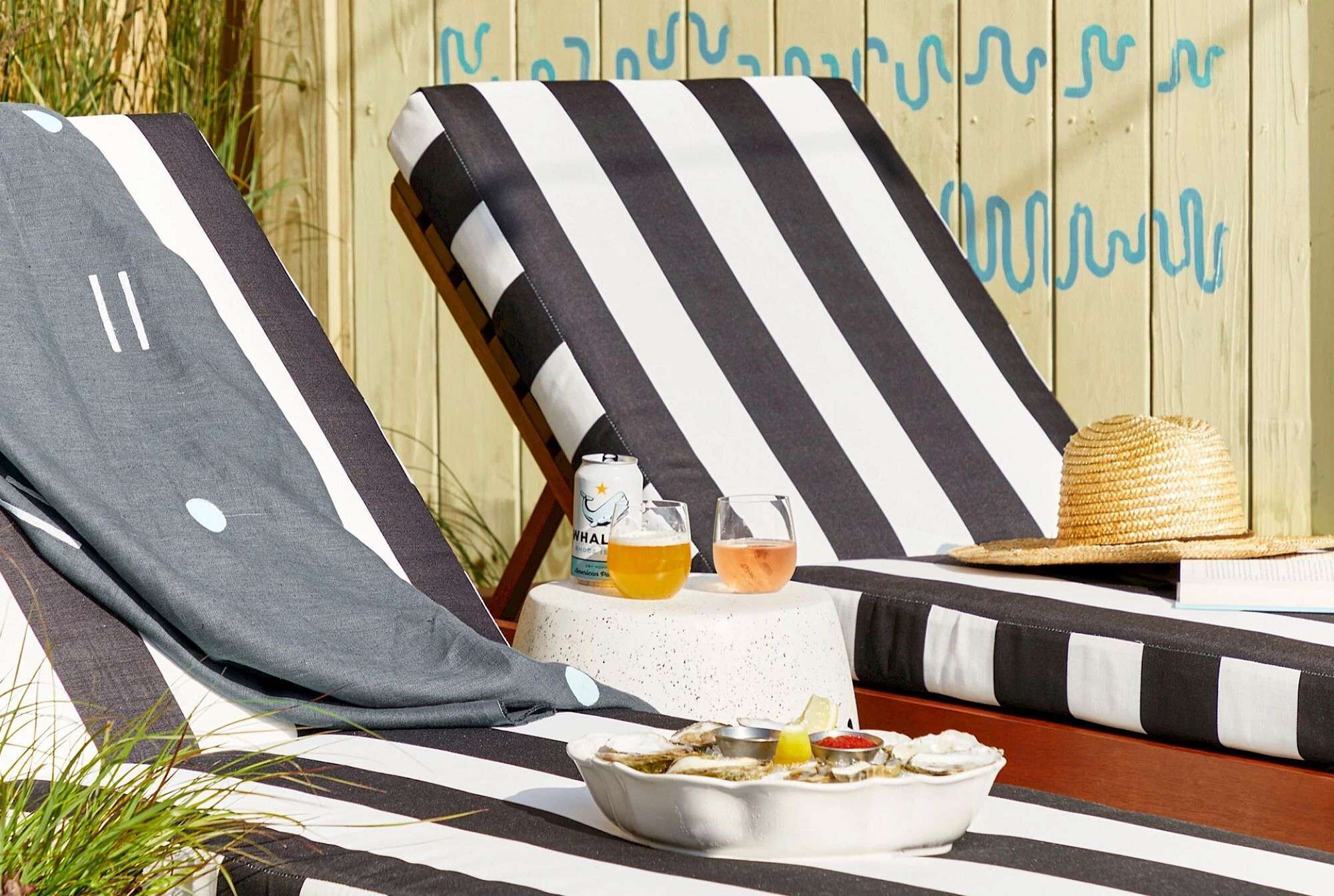 A beachside lounge with striped cushions, a small table with drinks and a basket, and a wooden backdrop with text.