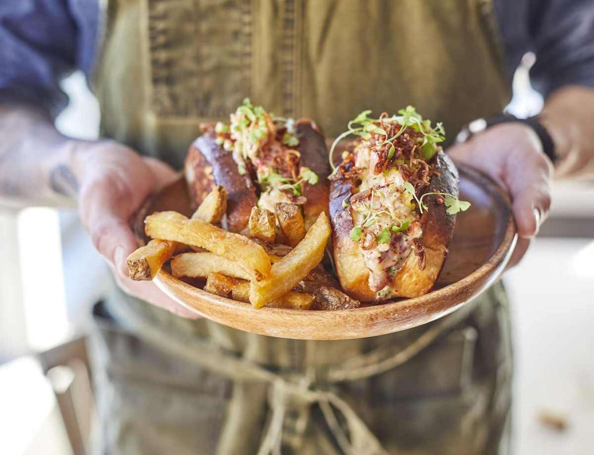 A person holding a wooden plate with two baked potatoes topped with a creamy sauce and crispy bacon, accompanied by a side of French fries.