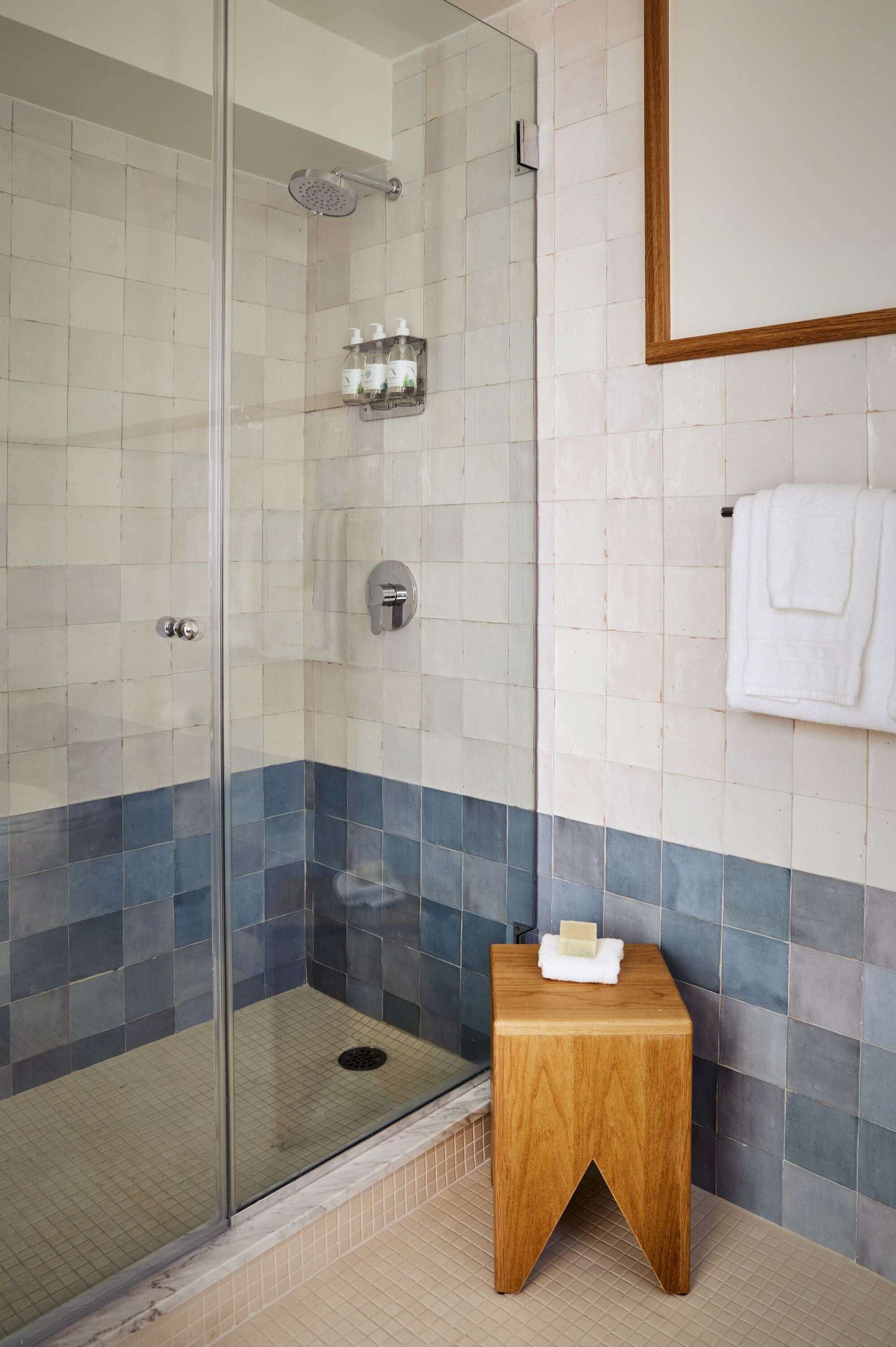 A glass-enclosed shower with a tiled floor, a wooden stool with a soap bar, and a towel hanging on the wall.