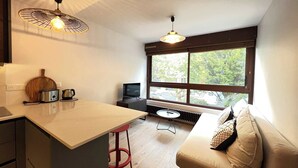 Cozinha compacta com torradeira, chaleira e tábua de corte de madeira. Há uma pequena mesa de jantar com um banquinho vermelho, um sofá com almofadas estampadas e uma TV. Janelas amplas oferecem vista para árvores.