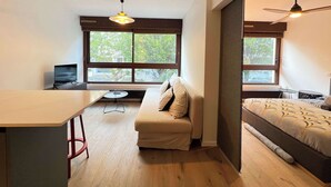 Quarto moderno com sofá, cama, mesa pequena, TV e grandes janelas com vista para árvores.