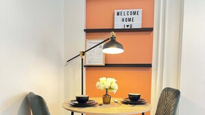 Un petit coin repas avec une table ronde, deux chaises, une lampe, un vase avec des fleurs et une pancarte « BIENVENUE À LA MAISON » accrochée au mur.
