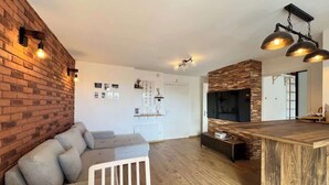 Ein modernes Wohnzimmer mit einer Ziegelwand, einem grauen Sofa, einem Holztisch und einer Küchenzeile inklusive großem Fernseher.