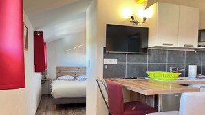 Een compacte hotelkamer met een bed, een kleine kitchenette, een eethoek met een tafel en stoelen, en een rood gordijn.
