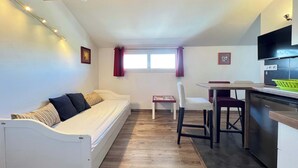 Een compacte kamer met een witte bank, een kleine eettafel en een kitchenette.