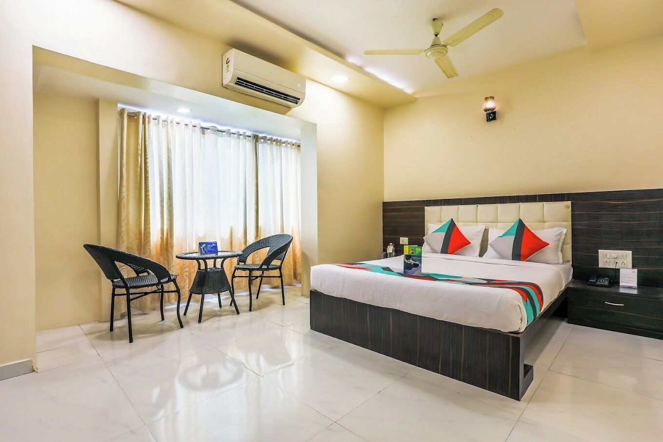 Habitación de hotel con cama, escritorio, dos sillas, aire acondicionado, ventilador de techo y ventana con cortinas.