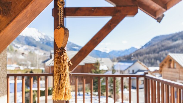 A wooden balcony with a rope hanging ornament, wooden railings, and a view of snow-covered houses and mountains in the background. - Bright Chalet with mountain view terrace - duplex (La Salle-les-Alpes)