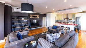 Una sala de estar moderna con un juego de sofá, una mesa de centro y un televisor. Hay una zona de cocina con una encimera roja y un refrigerador.