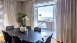 Comedor con una mesa y sillas, un jarrón con flores y vista a la playa a través de amplias ventanas.