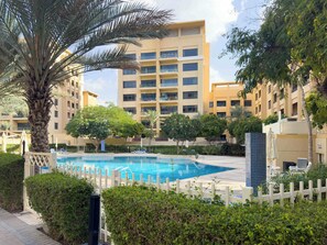 Une piscine entourée d’une clôture blanche, avec des palmiers et un immeuble à plusieurs étages en arrière-plan.