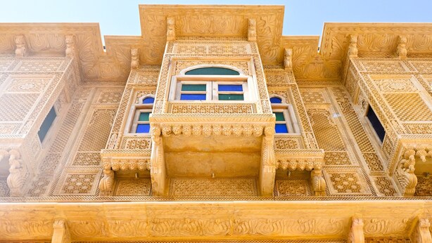 Un edificio con detalles arquitectónicos intrincados, incluyendo tallas ornamentales y un balcón con ventanas de tono azulado.