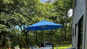 Een terras buiten met een tafel, stoelen, een blauwe parasol en een barbecue.