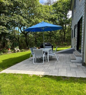 Eine Terrasse im Freien mit Tisch, Stühlen, einem blauen Sonnenschirm und einem Grill.